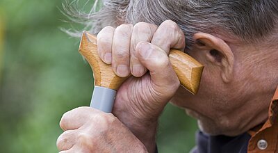 An elderly man holding a walking stick which he is leaning his head on.